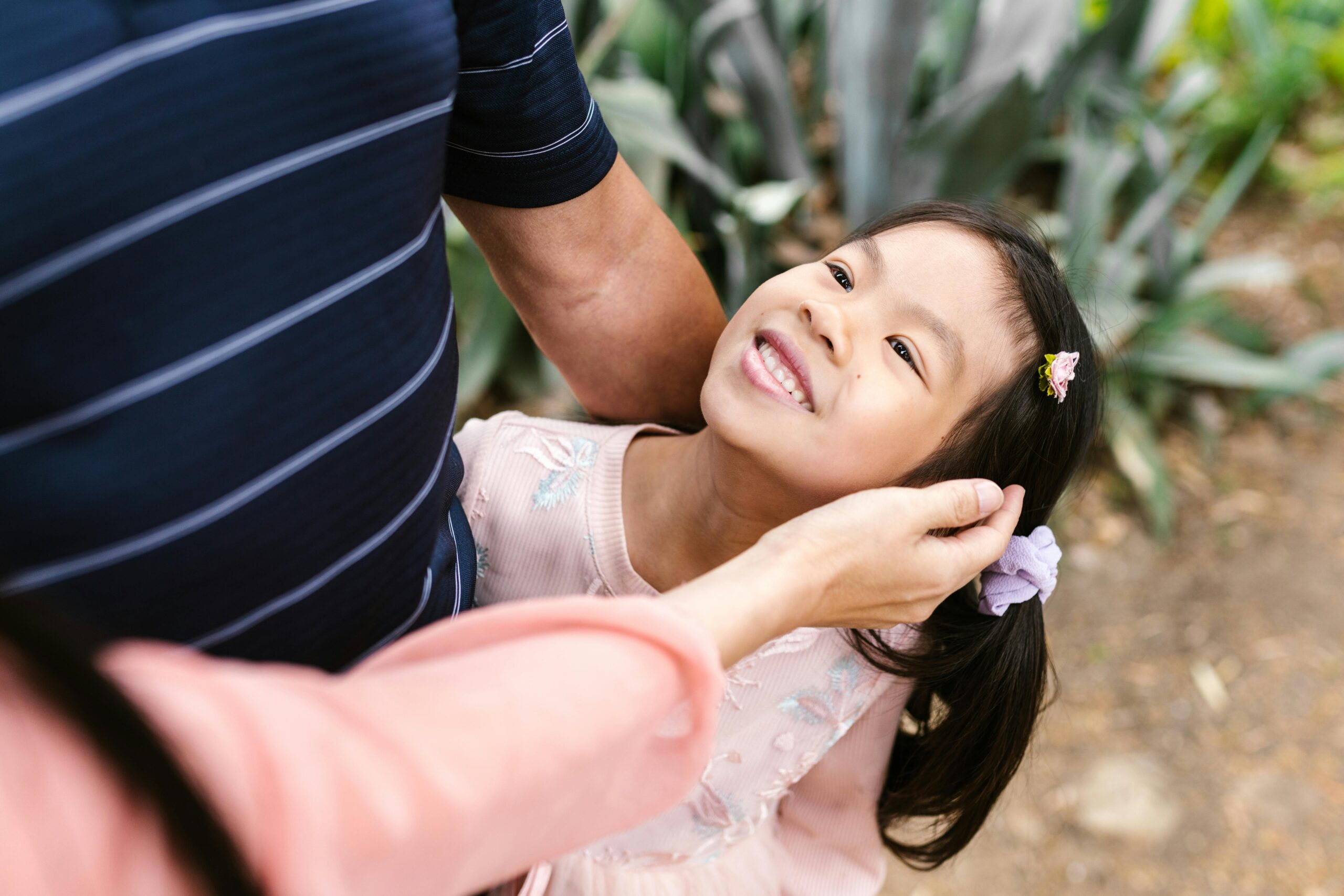 A joyful child smiling as a family member caresses her hair in an outdoor garden.